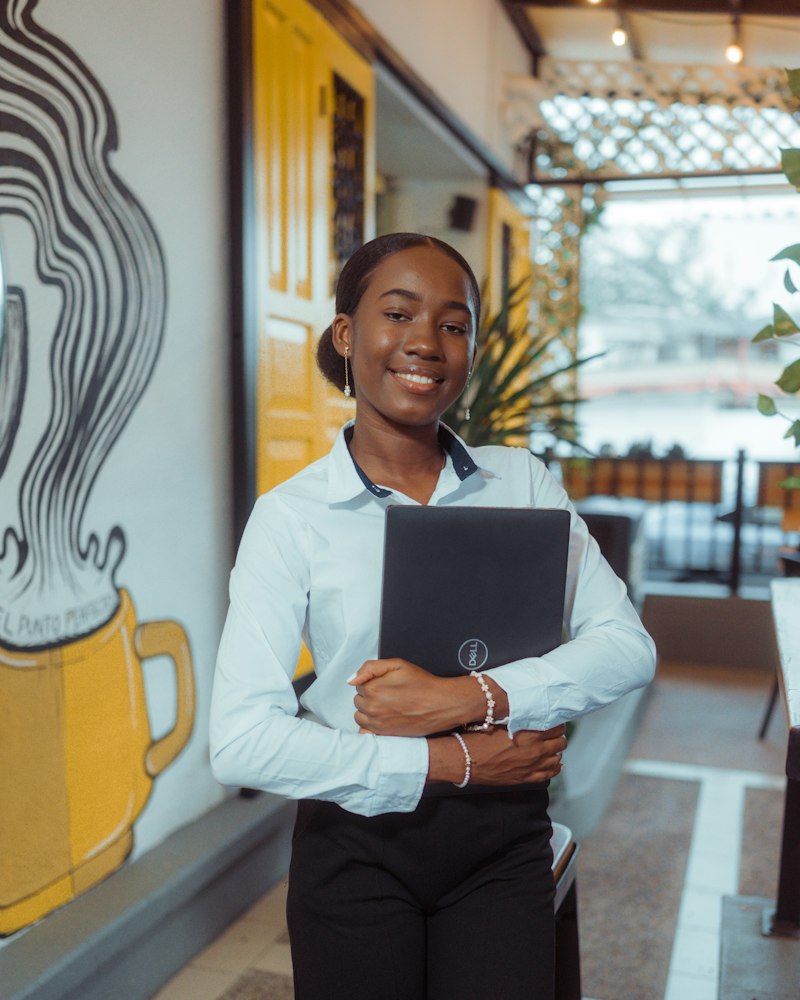 A smiling woman in a white shirt stands in a restaurant holding a laptop