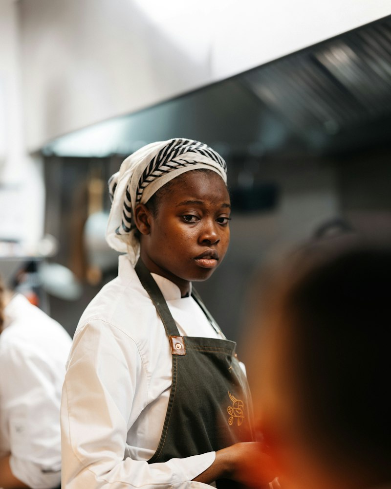 A young cook in kitchen whites and a head wrap listens closely in a restaurant kitchen