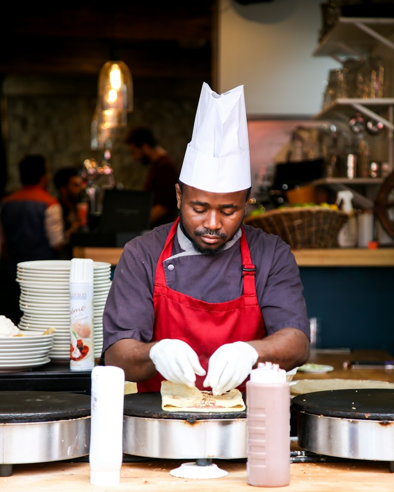 A cook in a chef’s hat and red apron prepares food at a crepe station