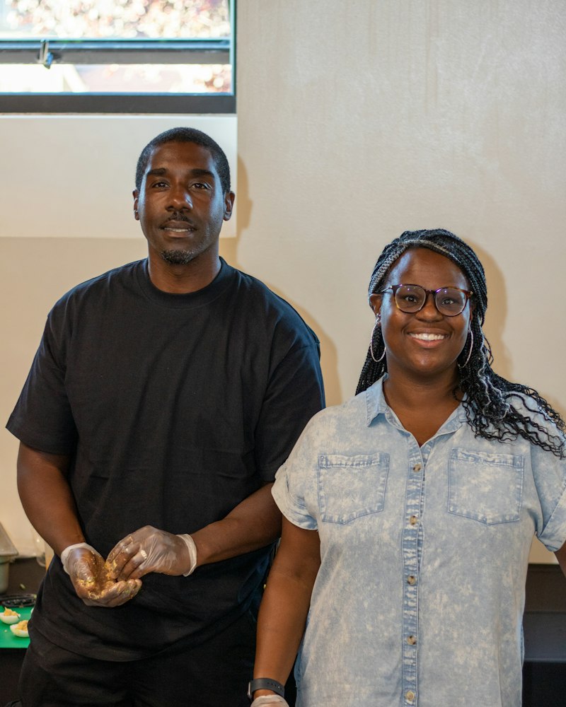 Two volunteers in gloves stand smiling behind a table of plated appetizers